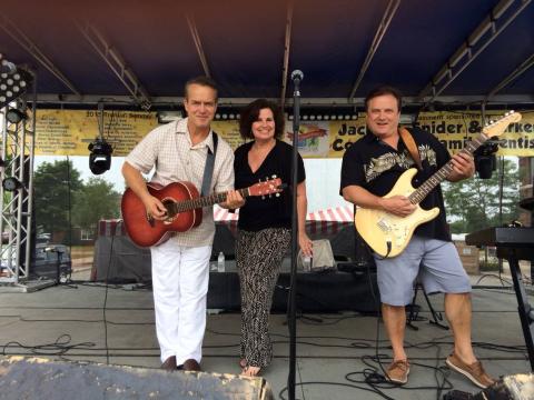 Two men and a woman on a stage, both men holding various types of guitars