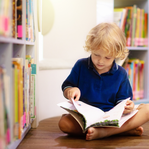 Child sitting on floor near library shelves reading a book