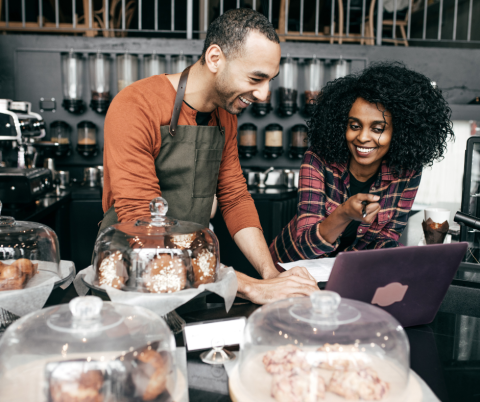 A man and woman behind a bakery counter, smiling and pointing to a computer screen.