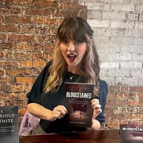 Photo of presenter, a white woman with brown hair, holding her book titled Bloodstained