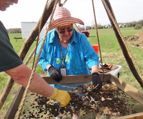 Person in a blue shirt, sunglasses, and a large hat sorting through a frame of soil at what appears to be an archaeological dig site.