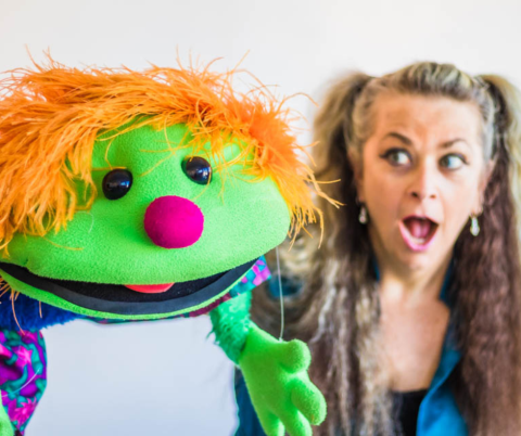 Green-skinned puppet with orange hair and a bright pink nose. Background blurry image of a woman with long blond hair and a surprised face.
