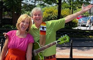 A man and a woman in bright clothes outdoors, smiling and happy. The man is holding a green guitar.