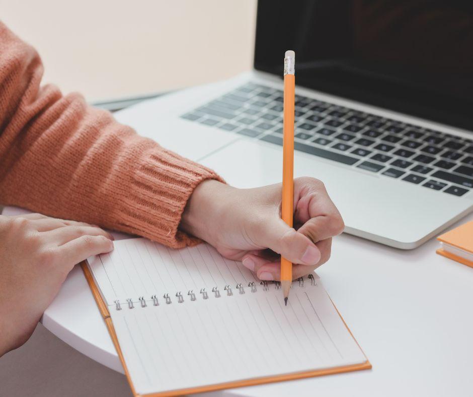 Person writing in a notebook with a pencil, laptop computer open behind them